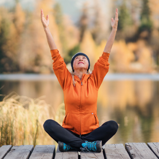 yoga-orange 1 Person sitting cross-legged on a wooden dock, arms raised in the air, facing a serene lake surrounded by autumn trees