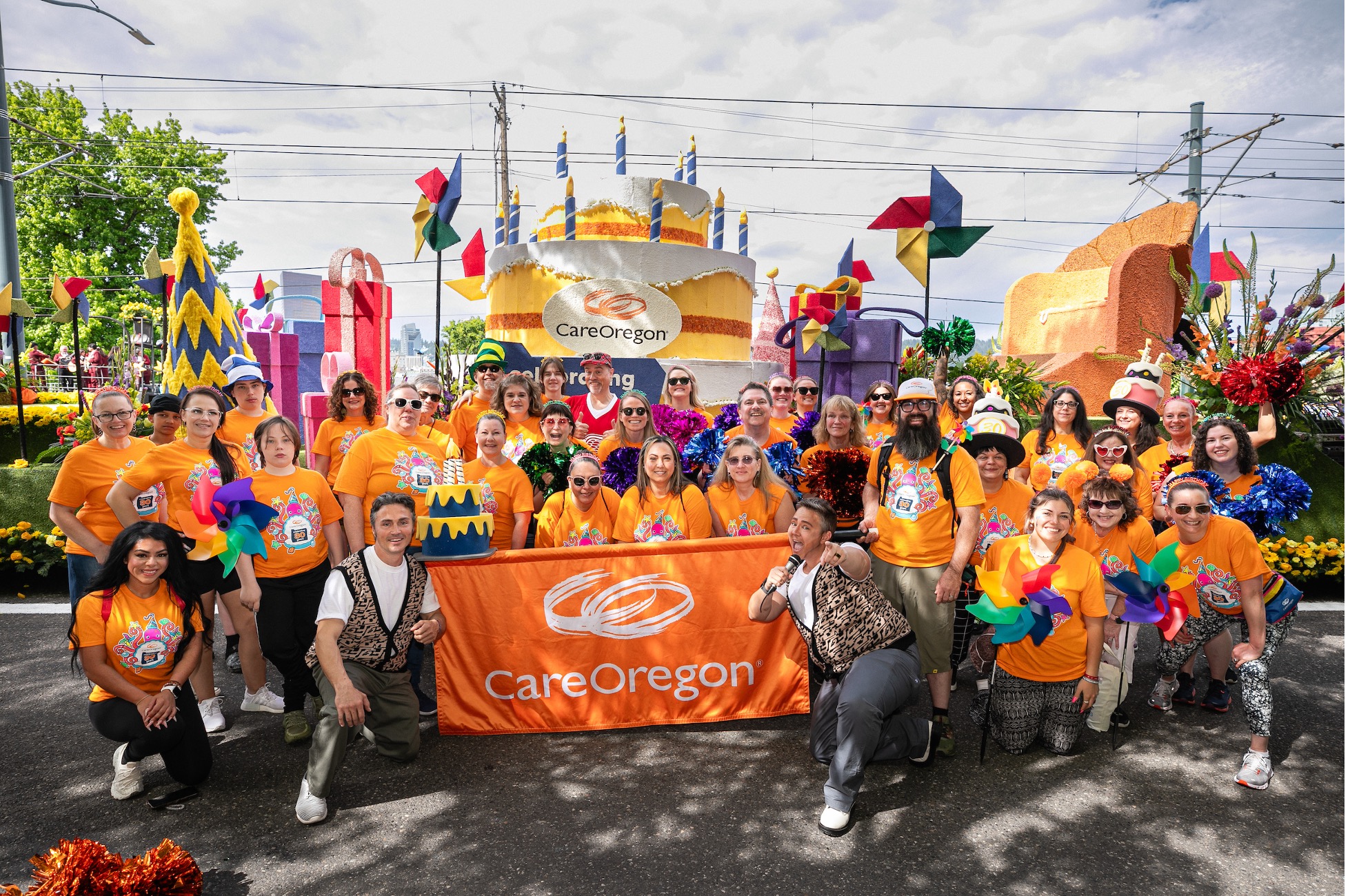 Un grupo de personas con camisetas naranjas de CareOregon reunidas frente a una carroza con un gran pastel de cumpleaños y un cartel que dice: "CarerOregon"