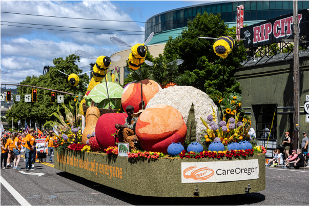 A colorful float adorned with large flowers and bees, sponsored by CareOregon, during a sunny parade