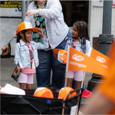 Two children receiving orange hats at an outdoor event, with an adult assisting them, in a street parade setting