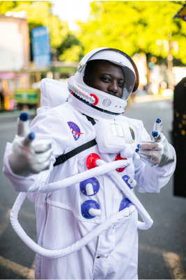 Starlight Parade_2023_JasonQuigley-1 A person in an astronaut costume standing on a city street, with their face obscured and a backdrop of blurred trees