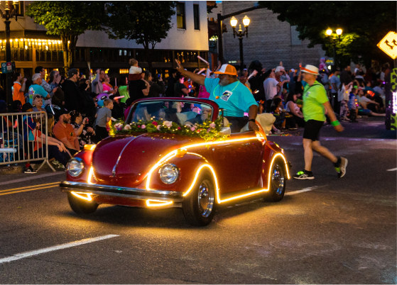 A vintage red car adorned with lights participating in a night parade, surrounded by spectators