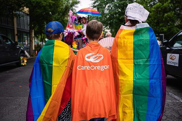 The backs of three boys are shown, two on the ends wearing rainbow capes and the one in the middle wearing an orange cape with the CareOregon logo centered