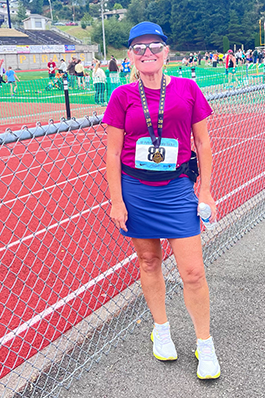 A woman in a fuscia shirt and blue running skirt, running shoes and sunglasses standing next to a red running track, smiling, holding a water bottle