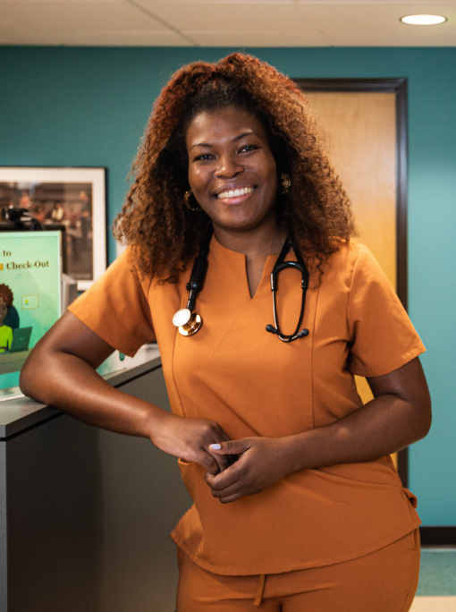 Dr. Gina Guillaume wears orange scrubs, a stethoscope and a big smile as she leans against the reception desk at North by Northeast Community Health Center, where she is medical director.