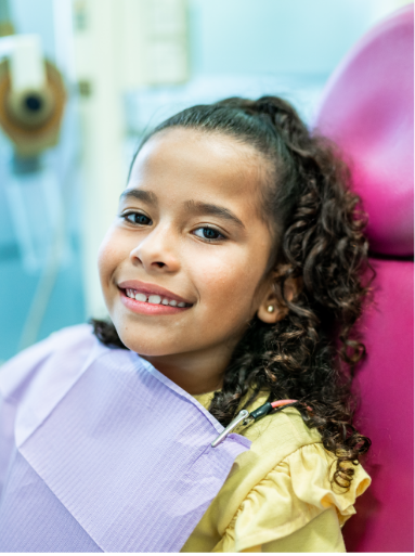 Una niña pequeña sonriendo a la cámara mientras está sentada en el sillón del dentista