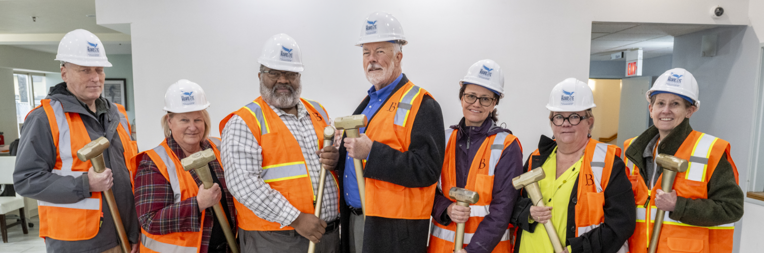 El personal de CareOregon y el equipo de construcción listos para comenzar la remodelación de Hawk's Eye Apartments, vestidos con chalecos de construcción de color naranja, cascos blancos y sosteniendo mazos.