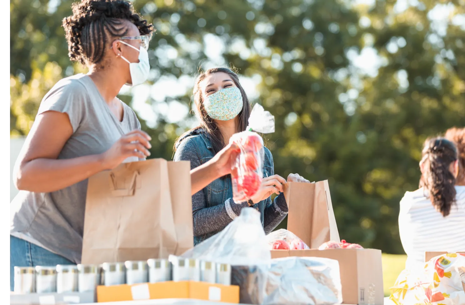 Two women wearing face covers, packing vegetables into paper bags in an outdoor setting