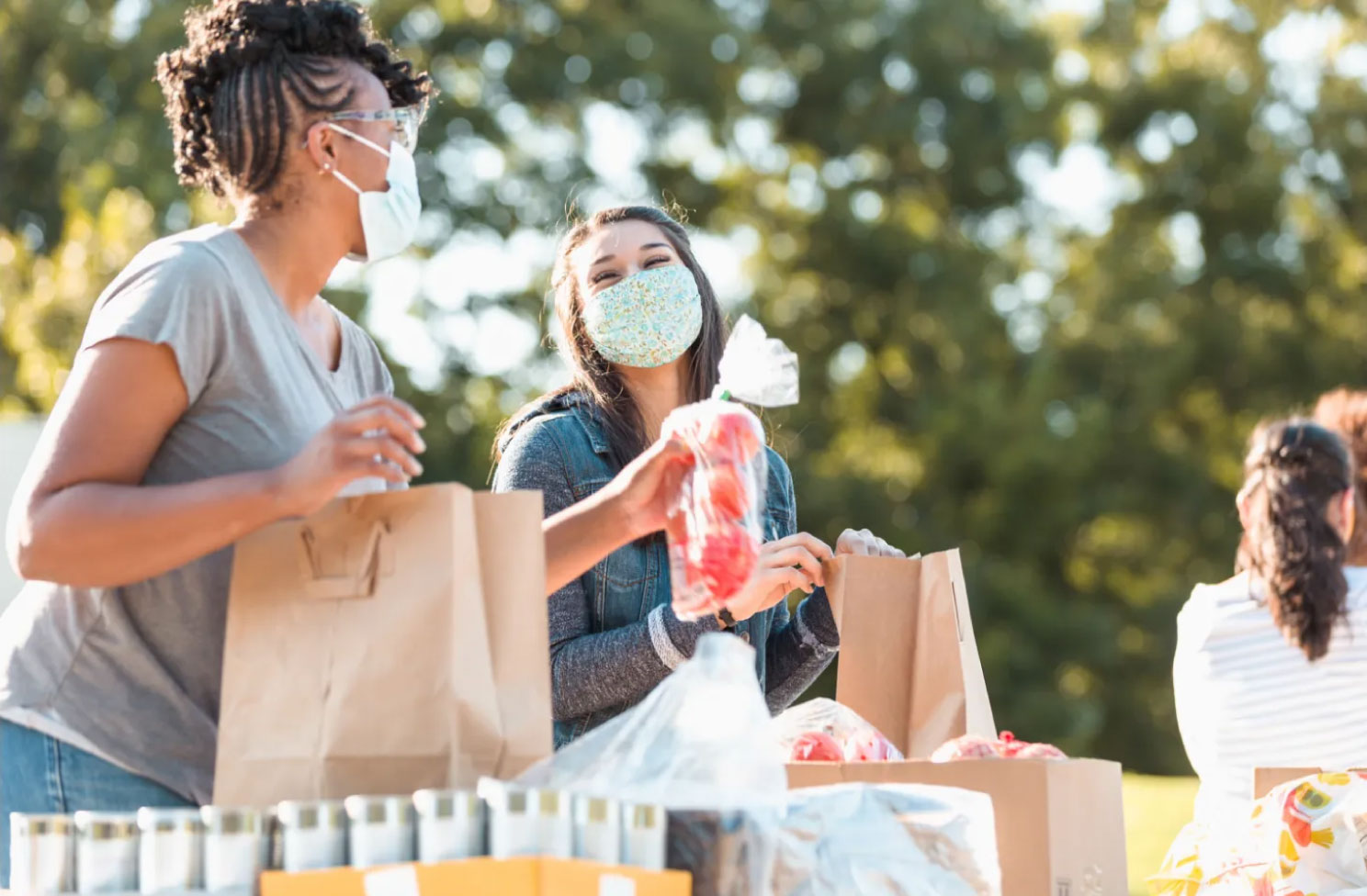 Two women wearing face covers, packing vegetables into paper bags in an outdoor setting