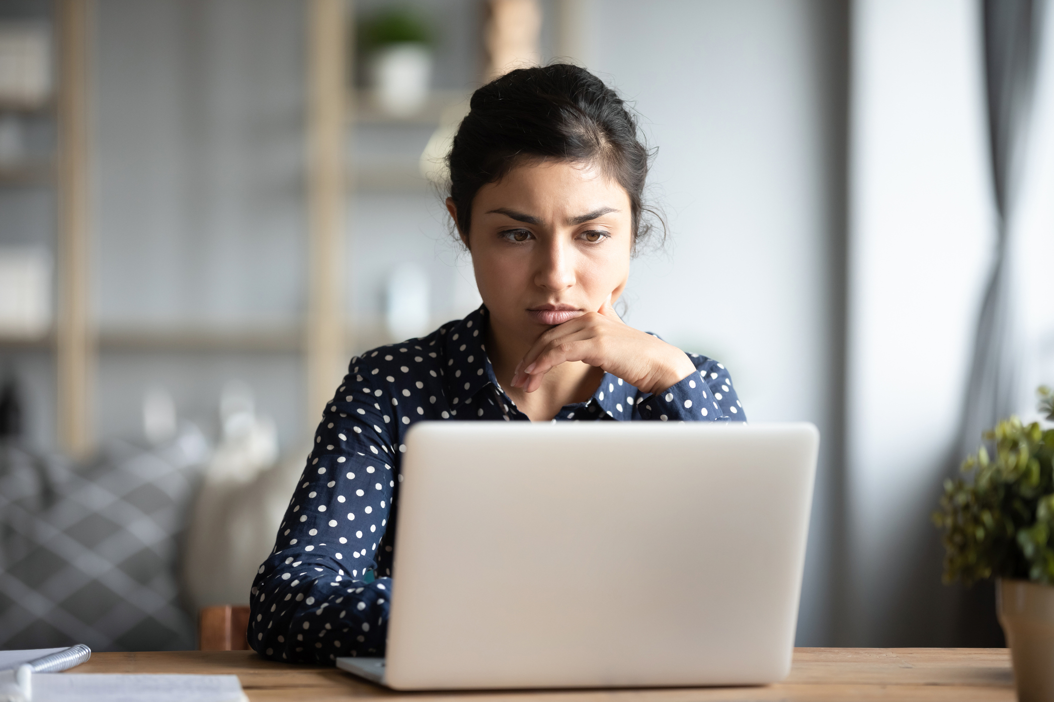 Woman with dark hair and blue polka dot shirt looks at laptop with a concerned look on her face.