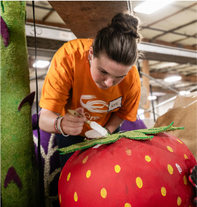 Person in an orange shirt painting details on a large, colorful strawberry art piece in a well-lit warehouse. 