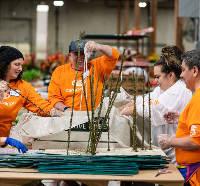Volunteers in orange shirts assembling green floral arrangements at a table in a warehouse setting 