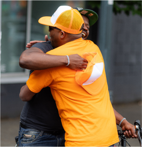Two people embracing in a hug, one wearing a bright orange t-shirt and the other in darker attire, with a glass building in the background. 