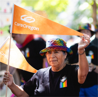 Person at an outdoor event waving a CareOregon flag, wearing a colorful hat and a black shirt with rainbow flag emblem. 
