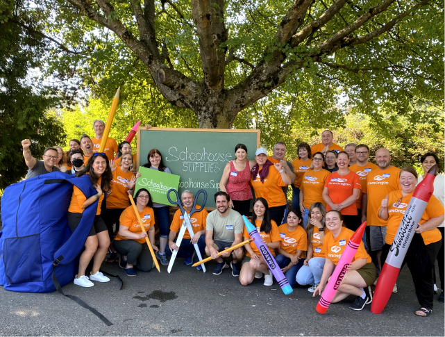 Un grupo de personas con camisetas naranjas de CareOregon paradas bajo un gran árbol, sosteniendo útiles escolares de gran tamaño