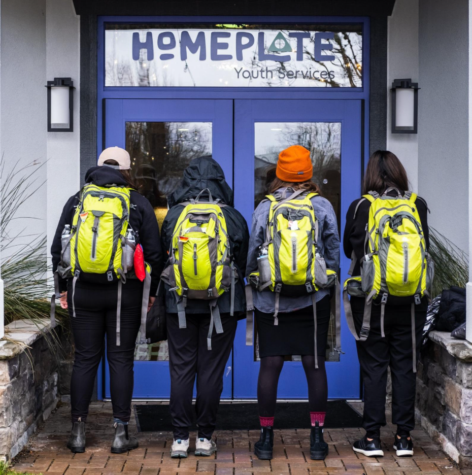 Four people with yellow backpacks stand in front of a door that reads Homeplate youth services
